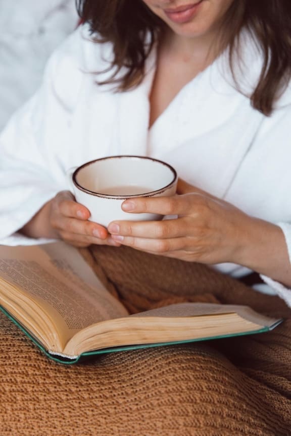 a person holding a hot drink while reading wrapped in a robe as part of anxiety calming and grounding techniques