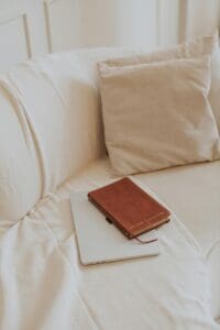 neutral space, well-lit, beige couch with pillows, closed laptop with brown leather journal/book 