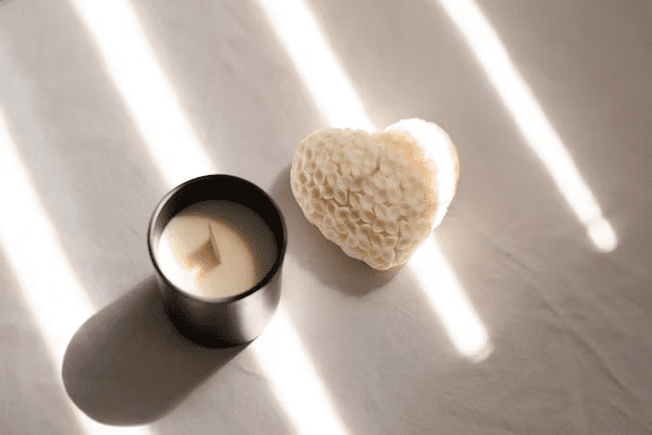 white background with light beams shining through from a window, black jarred candle that is not lit, white paper textured heart next to the candle