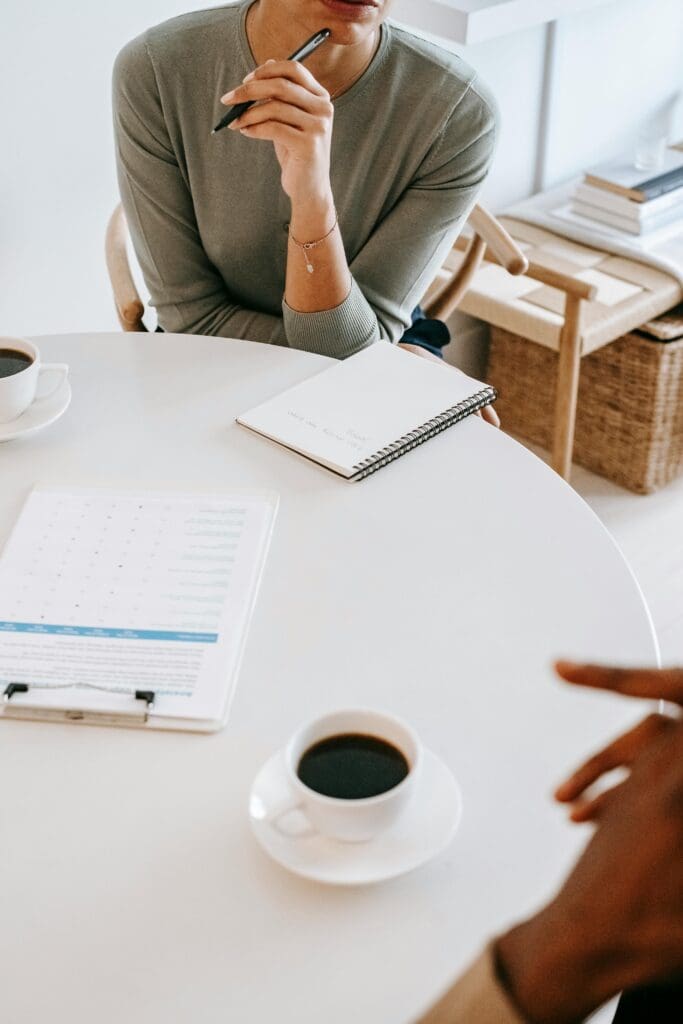 woman sitting at a white round table, notebook, clipboard, coffee cup on table, woman is sitting with her arm towards her chin holding a pen, listening to someone else talk