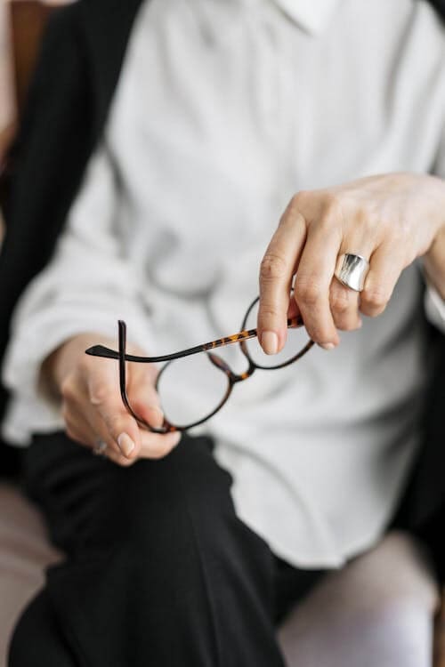 woman siting down, legs crossed, wearing a white blouse, holding her reading glasses in her hands as if she's listening