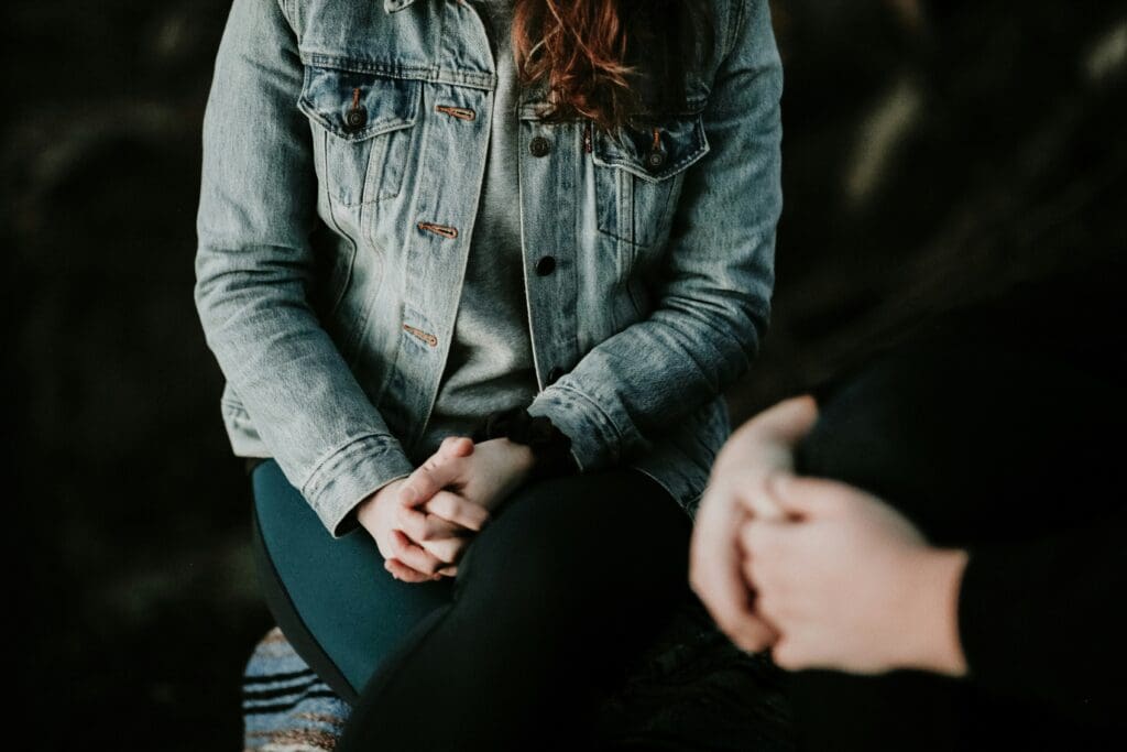 woman in jean jacket sitting on a bench hands folded on lap, with another person sitting on the bench with hands folded on their lap.