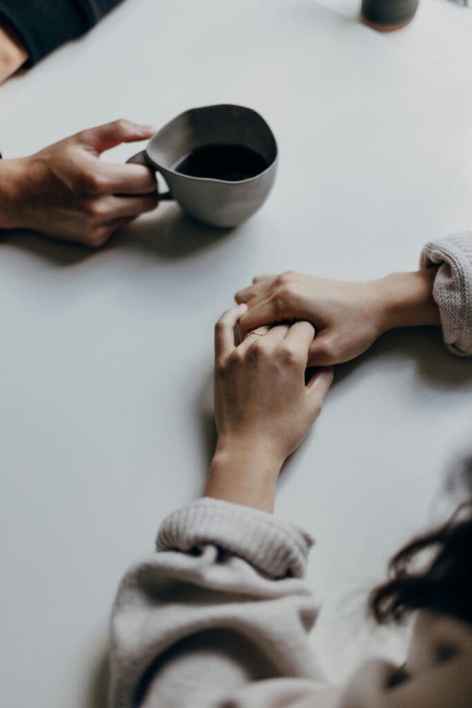 couple sitting across from each other at a table, woman is holding her hands together, man is holding a cup of coffee