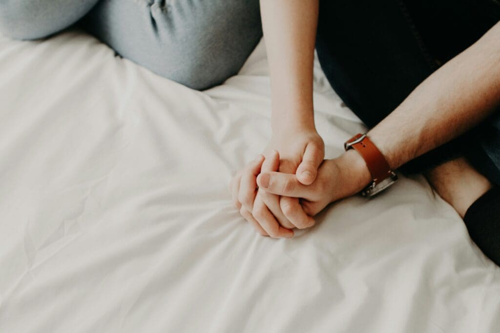 couple sitting together on the side of a bed, hands held together.
